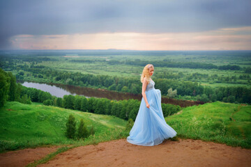 Fototapeta na wymiar pregnant woman in blue dress, green hills, river and sky. High quality photo