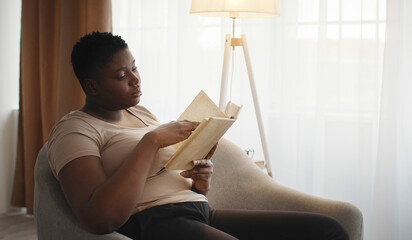 Self education concept. Young concentrated chubby african american lady reading interesting book,...
