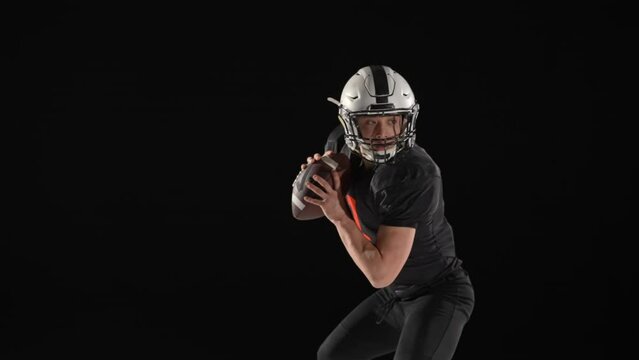 American football player wearing uniform, pads and helmet, throws the ball and scores a goal. Athlete rejoices in victory and points scored, posing against a black background. Slow motion.