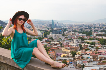 Fototapeta na wymiar A young girl dressed in a bright dress and hat, Sunglasses, poses against the backdrop of a view of the city. The concept of recreation and tourism in Georgia. Tbilisi. Space for text