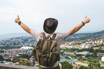 Back view of young man In a leather hat and retro backpack tourist on the background of panoramic view of Tbilisi city from high point, Georgia. Travel concept. Arms outstretched to the sides © Sergey