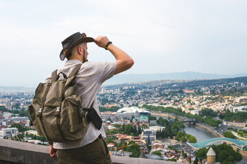 View of young man In a leather hat and retro backpack tourist on the background of panoramic view of Tbilisi city from high point, Georgia. Travel concept. Copy space © Sergey