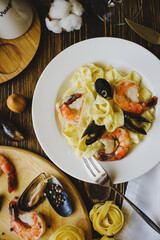 Photo of Italian pasta with mussels and shrimp in a restaurant on a textured wooden table and a dark background. Next to it is a round board with ingredients: mussels, cheese, sauces.