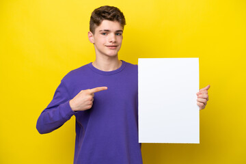 Teenager Russian man isolated on yellow background holding an empty placard with happy expression and pointing it