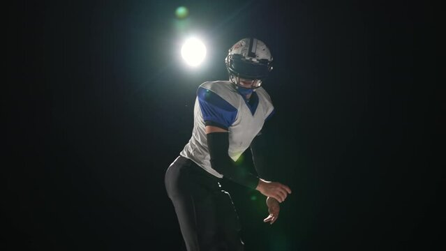 American football player wearing uniform, pads and helmet catches pass and runs on attack against black background with spotlight. Players run attacking to score touchdown points. Slow motion.