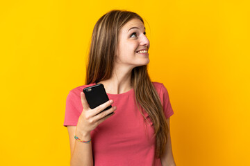 Young woman using mobile phone isolated on yellow background looking up while smiling
