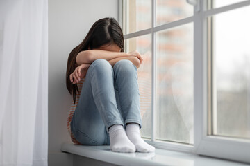 Upset asian girl schooler sitting on windowsill and crying