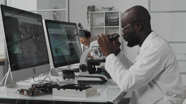 African American engineer in lab coat sitting at desk with two computer screens and examining chip with microscope
