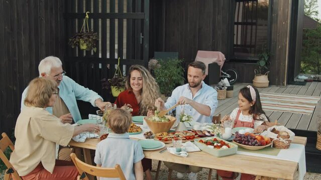 Medium of Caucasian family members of three generations sitting together at table in backyard of summer house on sunny weekend, having healthy lunch, smiling and chatting