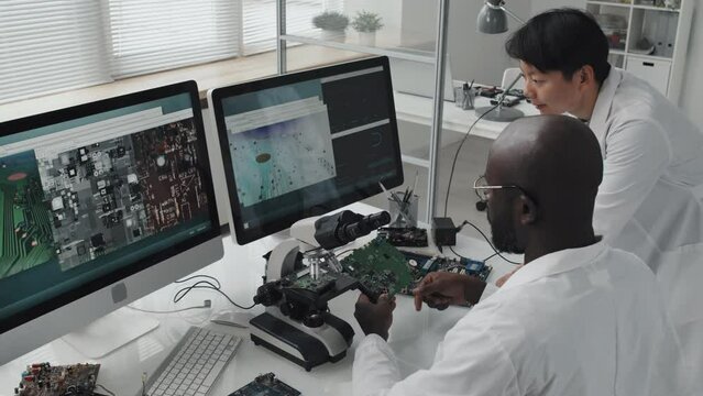High angle shot of African American male engineer discussing circuit board and its macro images on computer screens with Asian colleague while cooperating in laboratory
