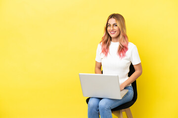 Naklejka premium Young woman sitting on a chair with laptop over isolated yellow background posing with arms at hip and smiling