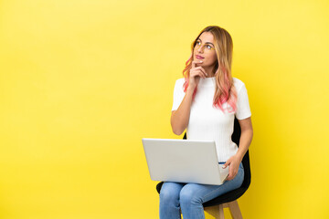 Young woman sitting on a chair with laptop over isolated yellow background looking up while smiling