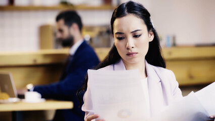 focused asian businesswoman looking at documents near blurred businessman