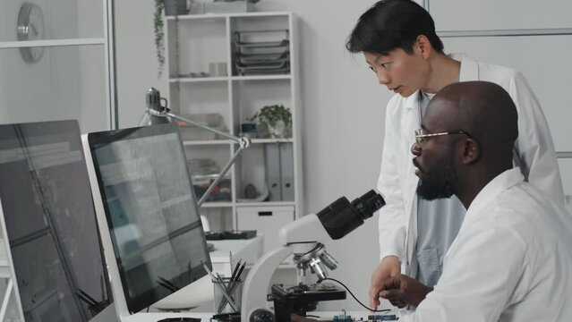 Tilt up shot of African American male engineer and his Asian female colleague discussing image of electronic chip on computer and having discussion while doing lab research with microscope