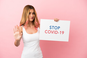 Young woman over isolated pink background holding a placard with text Stop Covid 19 and doing stop...