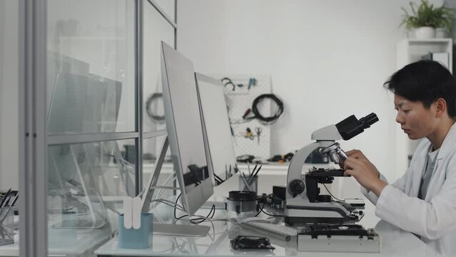 Tracking shot of Asian female lab technician using microscope while African American electronics engineer working with multimeter and computer at desk