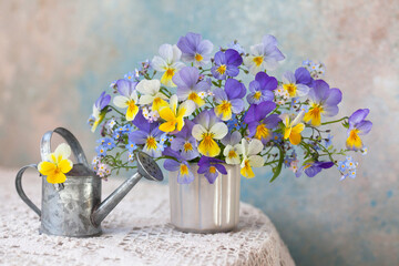 A bouquet of pansies viola and forget-me-not flowers in a vase and a watering can on a table with a tablecloth against the background of a colored wall. Romantic postcard, blur, selective focus. - 486472518