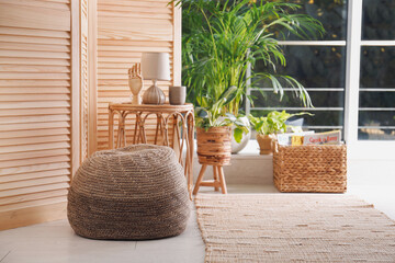 Stylish knitted pouf, wicker furniture and beautiful houseplants in room. Interior design