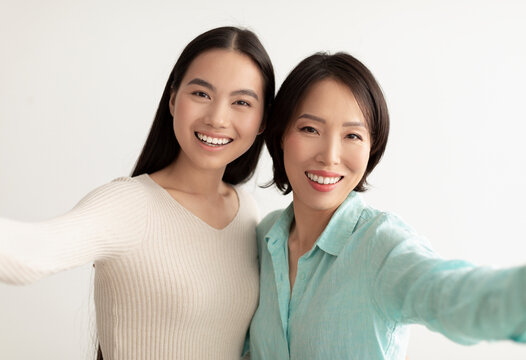 Happy young Asian woman and her mature mother taking selfie together on white studio background