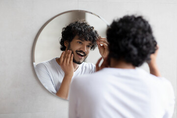 Male beauty and haircare routine. Handsome indian man touching beard and hair, looking in mirror in...
