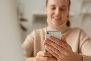 Closeup portrait of woman wearing beige sweater posing with cell phone in hands, looking smiling at devise display, communicating online, watching funny videos.