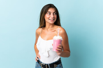 Young woman with strawberry milkshake isolated on blue background posing with arms at hip and...