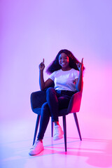 Smiling young black woman sitting in armchair, pointing upwards, offering blank space in neon light, mockup for ad