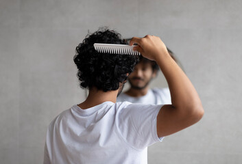 Back view of indian curly man combing hair standing near mirror in bathroom. Male haircare routine...