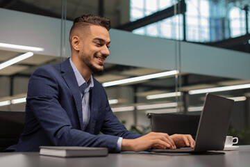 Cheerful middle eastern businessman typing on notebook, office interior