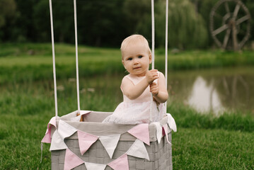A beautiful little girl in a basket with a balloon and a cake celebrates her first birthday