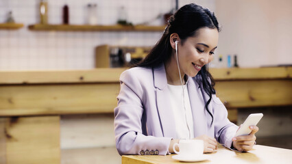 smiling asian woman in suit listening music in earphones and using smartphone near cup of coffee in cafe