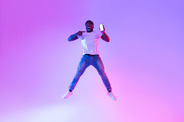 Fototapeta na wymiar Full length of excited young black guy jumping with cellphone, pointing at empty screen in neon light, mockup