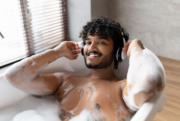 Calm indian man in wireless headphones lying in hot bubbly bath and enjoying his playlist in bathroom at home