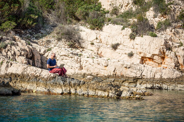 Digitaler Nomade mit Sonnenbrille und roter Hose sitzt auf Felsen am Meer und arbeitet am Laptop
