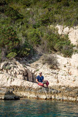 Digitaler Nomade mit Laptop, Sonnenbrille und roter Hose sitzt auf Felsen am Meer und blickt in die Ferne, Totale, Hochformat