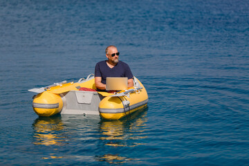 Digitaler Nomade mit Sonnenbrille sitzt in gelbem Gummiboot am Meer und arbeitet am Laptop, Nahaufnahme