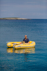 Digitaler Nomade mit Sonnenbrille sitzt in gelbem Gummiboot am Meer und arbeitet am Laptop, Hochformat