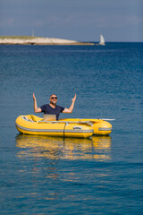 Digitaler Nomade mit Sonnenbrille und Laptop sitzt in gelbem Gummiboot am Meer und jubelt, Hochformat