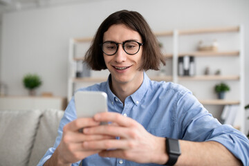 Positive young man sitting on couch with smartphone, chatting with ladies, using dating mobile application on cellphone