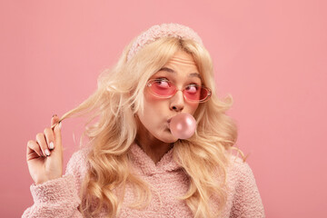 Pretty blonde lady blowing bubble from chewing gum and twisting lock of hair on her finger, pink background