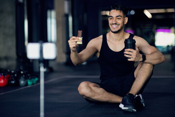 Fototapeta na wymiar Sport Snacks. Handsome Arab Guy Holding Protein Bar And Fitness Shaker Bottle