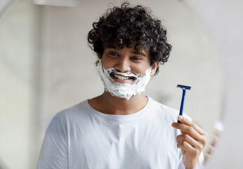Cheerful indian man with face covered with shaving foam holding safety razor while looking at...