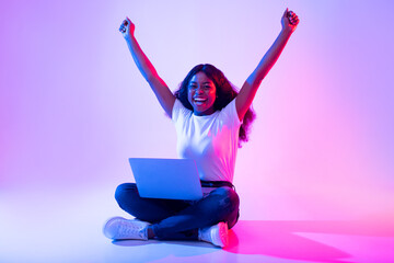 Overjoyed young black female with laptop raising arms up, celebrating achievement or success in...