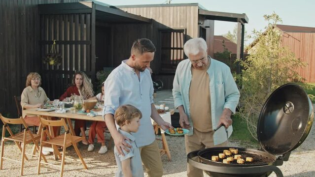 Medium long of white-haired grandpa putting hot corn from grill onto plate for adult son and little boy, standing in backyard of house on sunny summer day, talking