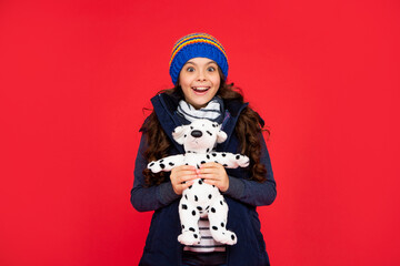 surprised child in puffer jacket hold toy. teen girl on red background. happy childhood.