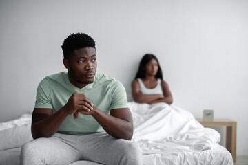 Depressed sad young black man ignores offended lady after quarrel sits on bed in bedroom interior, copy space