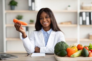 Cheerful african american female dietitian holding carrot at medical clinic, sitting at workplace...