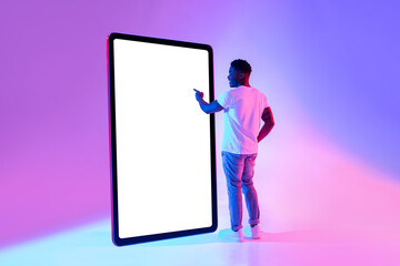 Happy young African American man standing near big tablet computer with mockup, touching screen in...