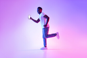 Fototapeta na wymiar Excited young black guy with smartphone running in neon light, showing mockup for design on mobile device screen
