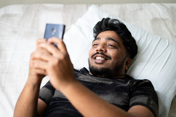 happy young man chatting on mobile phone while sleeping on bed - concept of using social media,dating app, flirting and technology.